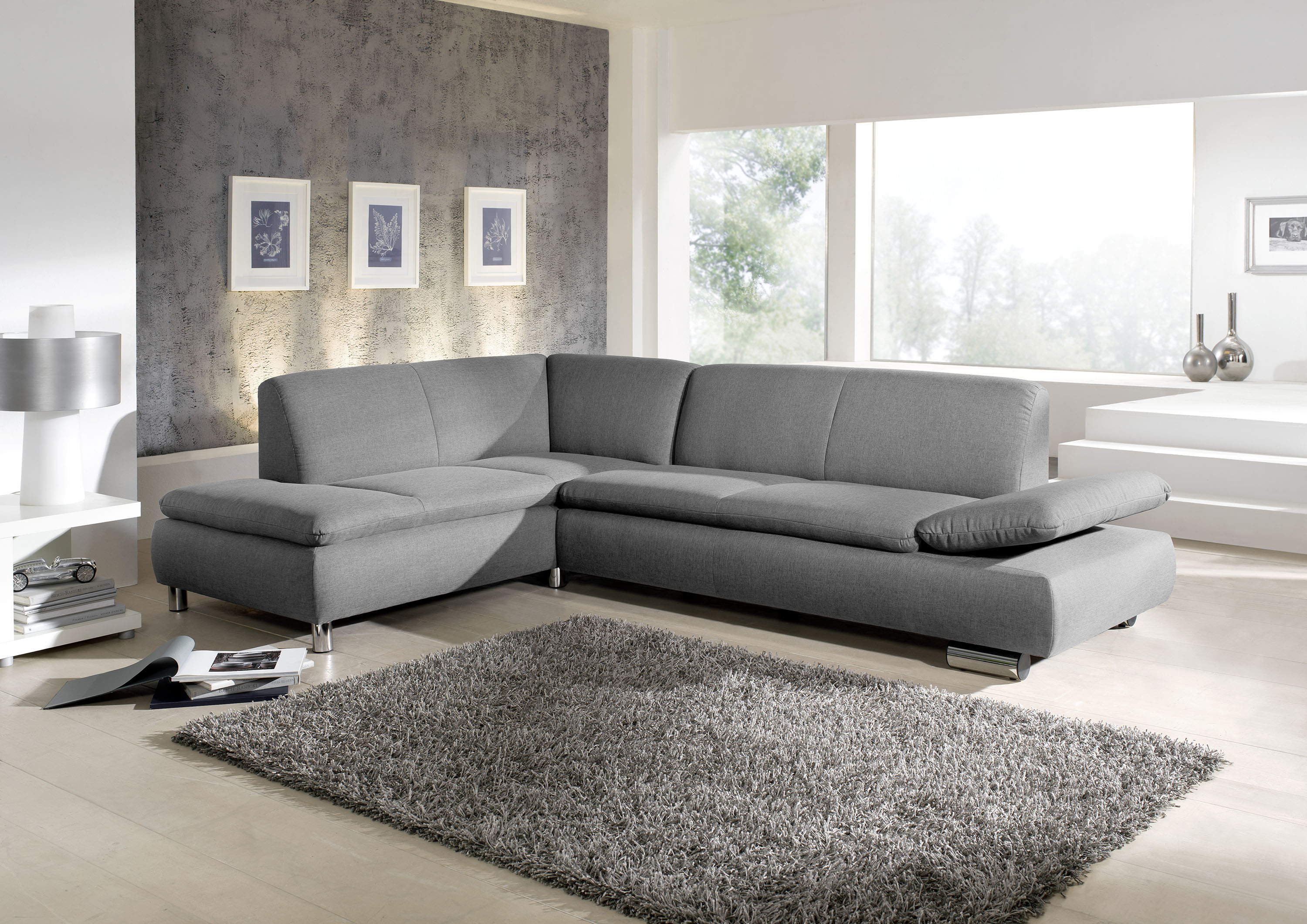 graues ecksofa mit hochgeklappter armlehne und verchromten metallfüssen in einem hellen wohnzimmer mit grauen teppich